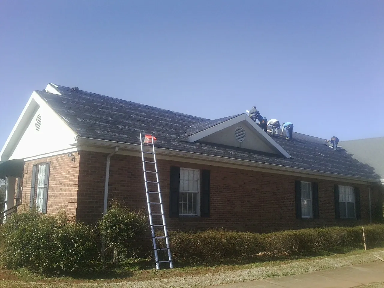 Crew tearing shingles off a brick ranch roof with a ladder set — Metal Roof Repair work by our crew in Gleason
