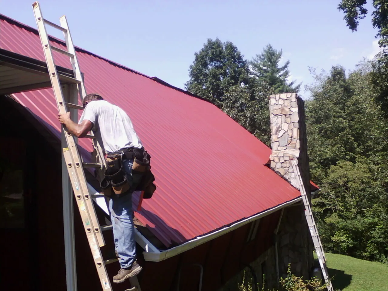 Roofer on a ladder beside a red metal roof and stone chimney — Soffit & Fascia Repair by Roofing Brothers in Gleason